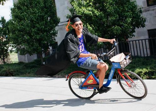 A graduate in cap and gown riding a KU bicycle
